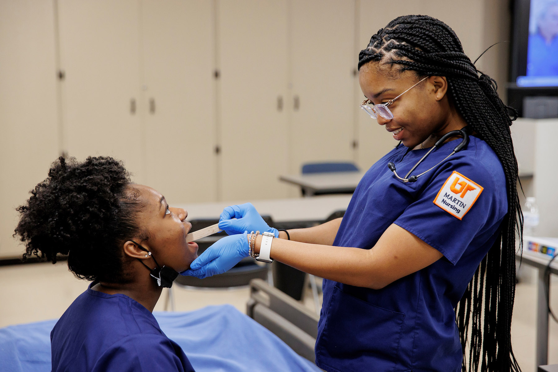 Medical professional in blue scrubs examines patient's throat, wearing protective gloves in clinical setting with cabinets in background.