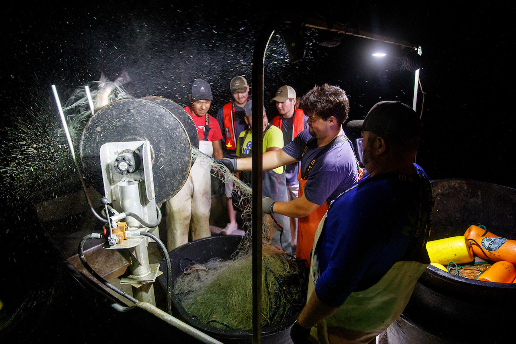 Night fishing crew processing catch on boat, working around large machine with nets and bright work lights in misty environment.